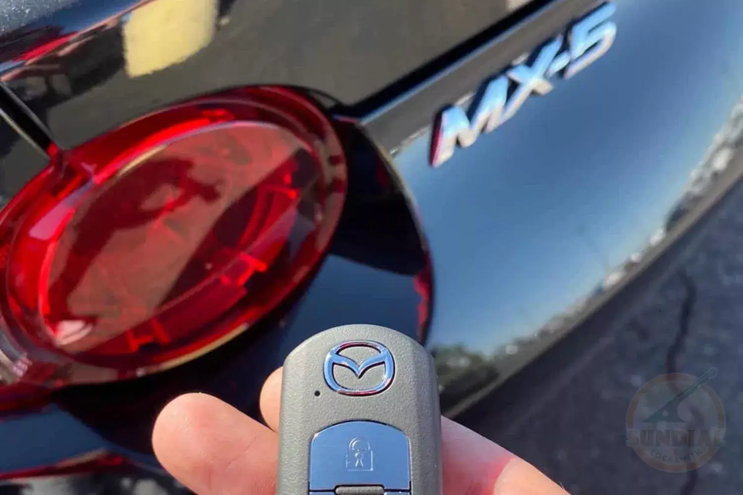MAZDA MX-5 KEY REPLACEMENT A hand holding a Mazda smart key fob in front of the rear of a black Mazda MX-5 vehicle.