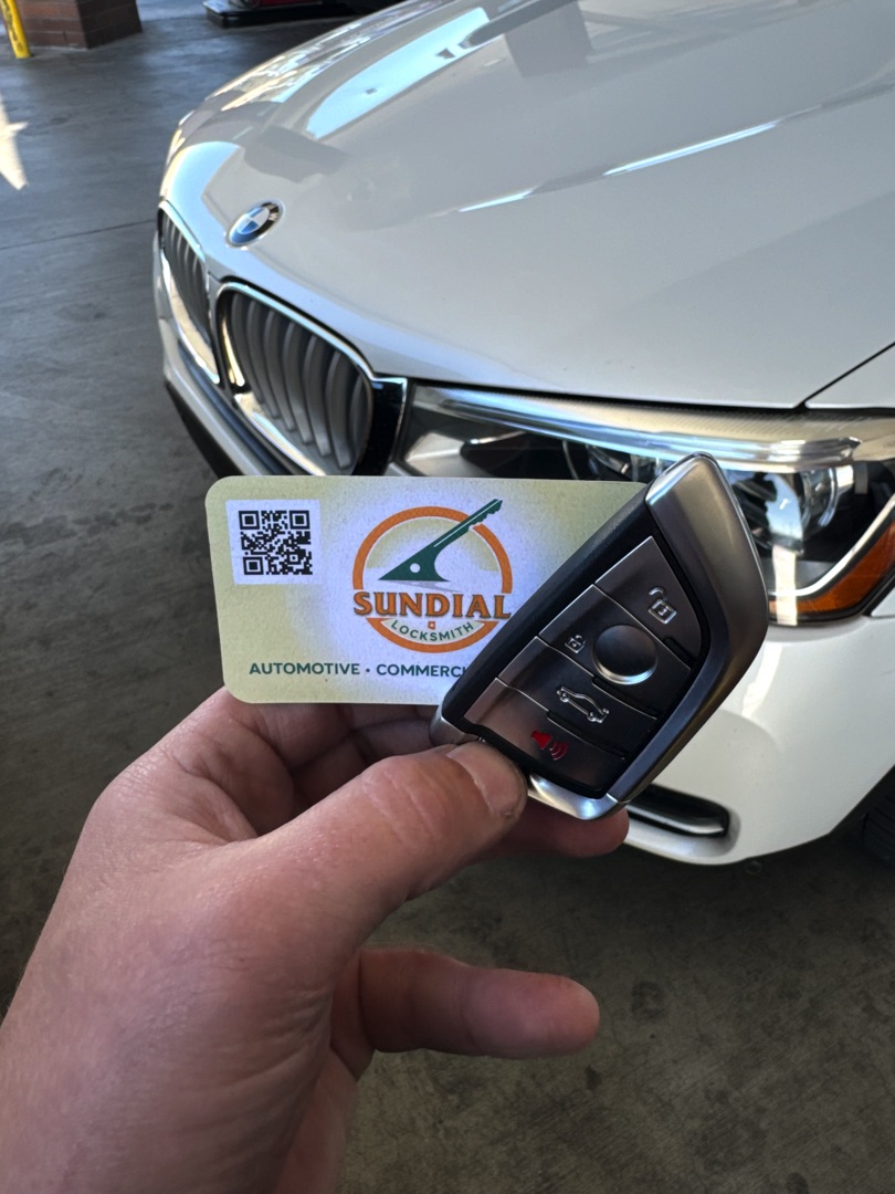 Why High-Rise Buildings Need a Different Kind of Locksmith A hand holding a BMW smart key fob and a Sundial Automotive Locksmith business card in front of a white BMW vehicle.