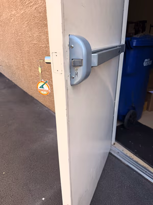 Panic Bar Installation: What Every Business Owner Needs to Know A gray metal push bar panic exit device installed on a white commercial door with a cable attachment, viewed from the exterior side.