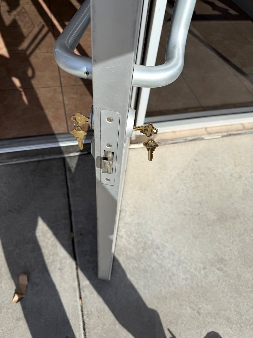 How to Secure a Sliding Glass Door That a Burglar Can't Just Lift Off Close-up of a sliding glass door edge showing two brass keyed lock cylinders installed in a silver aluminum door frame.