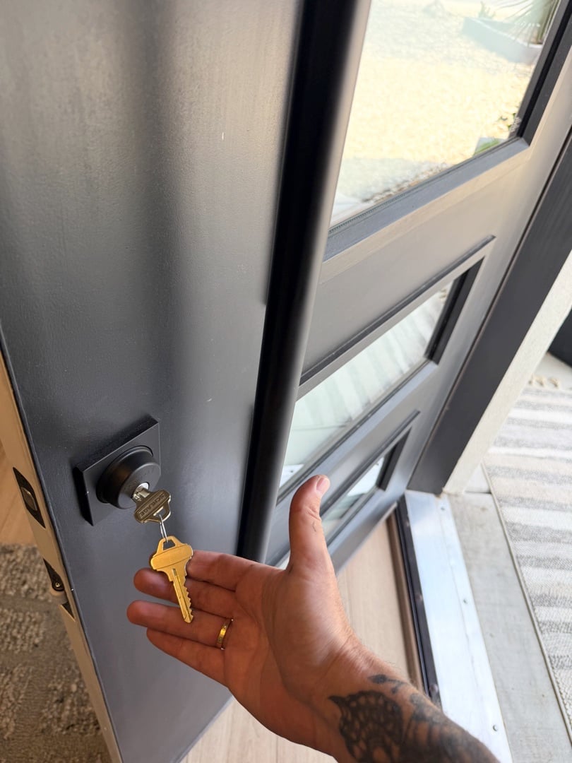 Lost Your House Keys? Here's What to Do Before You Panic A tattooed hand holds a yellow key near the lock of a dark gray front door with a glass panel.