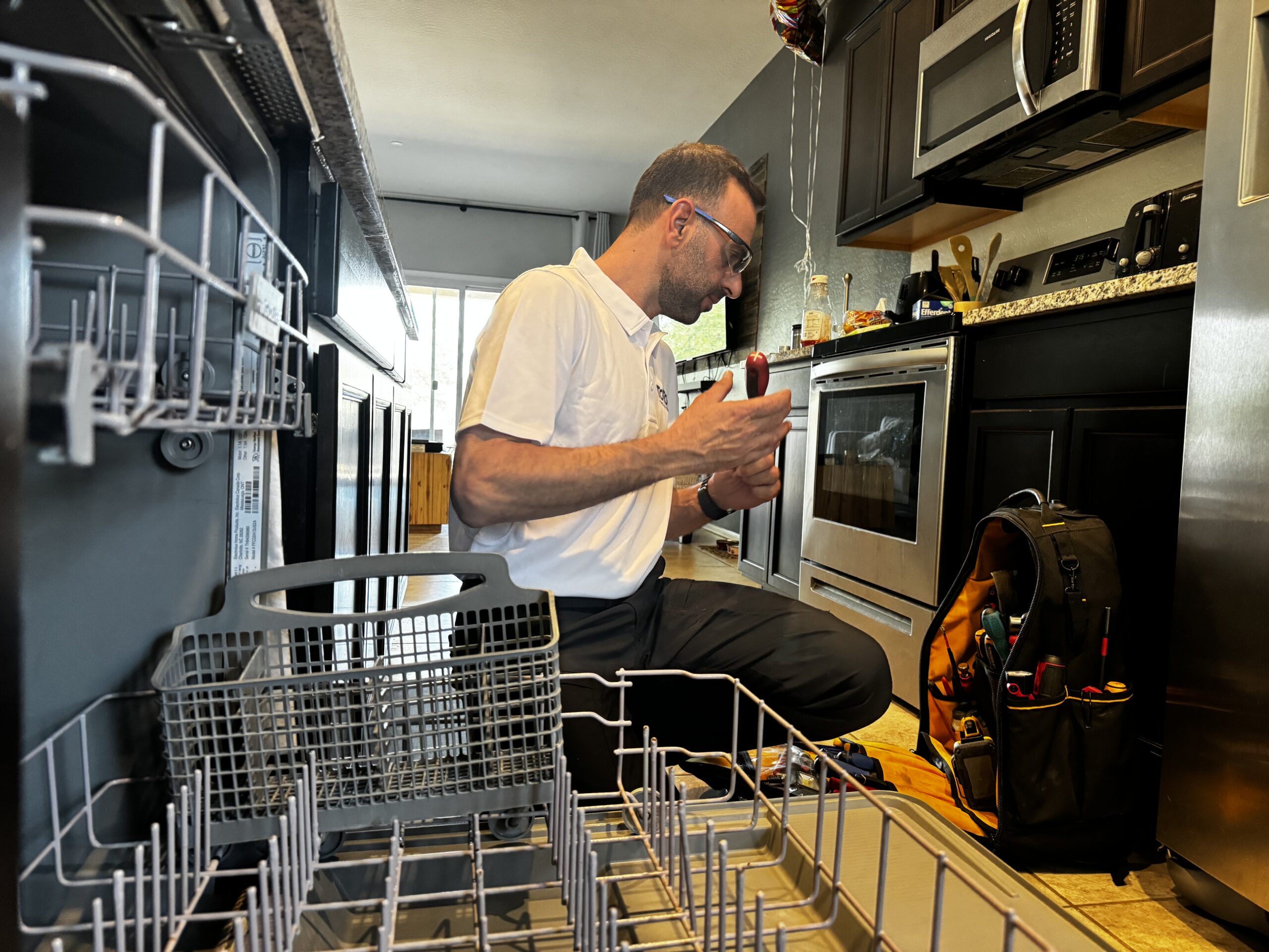 private-party-02 A technician kneels in a kitchen, examining a tool by an open dishwasher, with a tool bag nearby and kitchen appliances in the background.