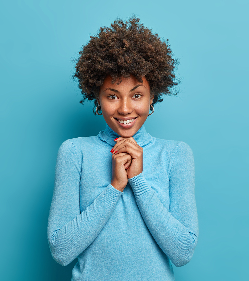 Portrait of happy girl has Afro hair keeps hands under chin looks with pleased satisfied expression at camera hears wonderful news dressed in casual turtleneck isolated on blue wall. Face expressions