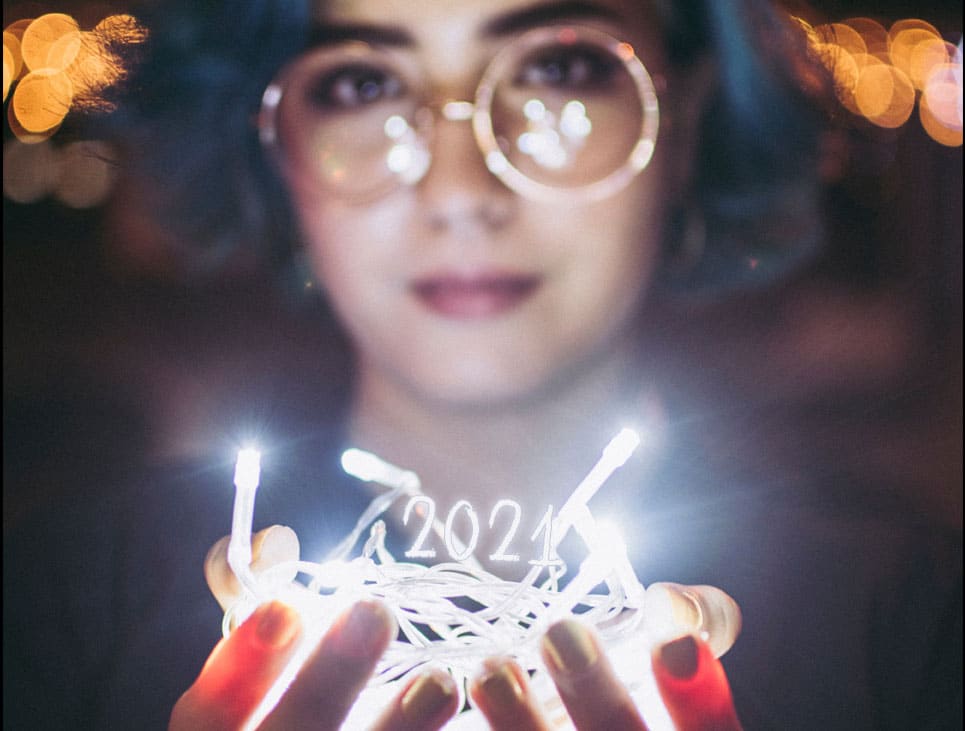 Photographie d'une femme aux cheveux bleus tenant une guirlande de lumières dans ses mains, surmontée des chiffres "2021"