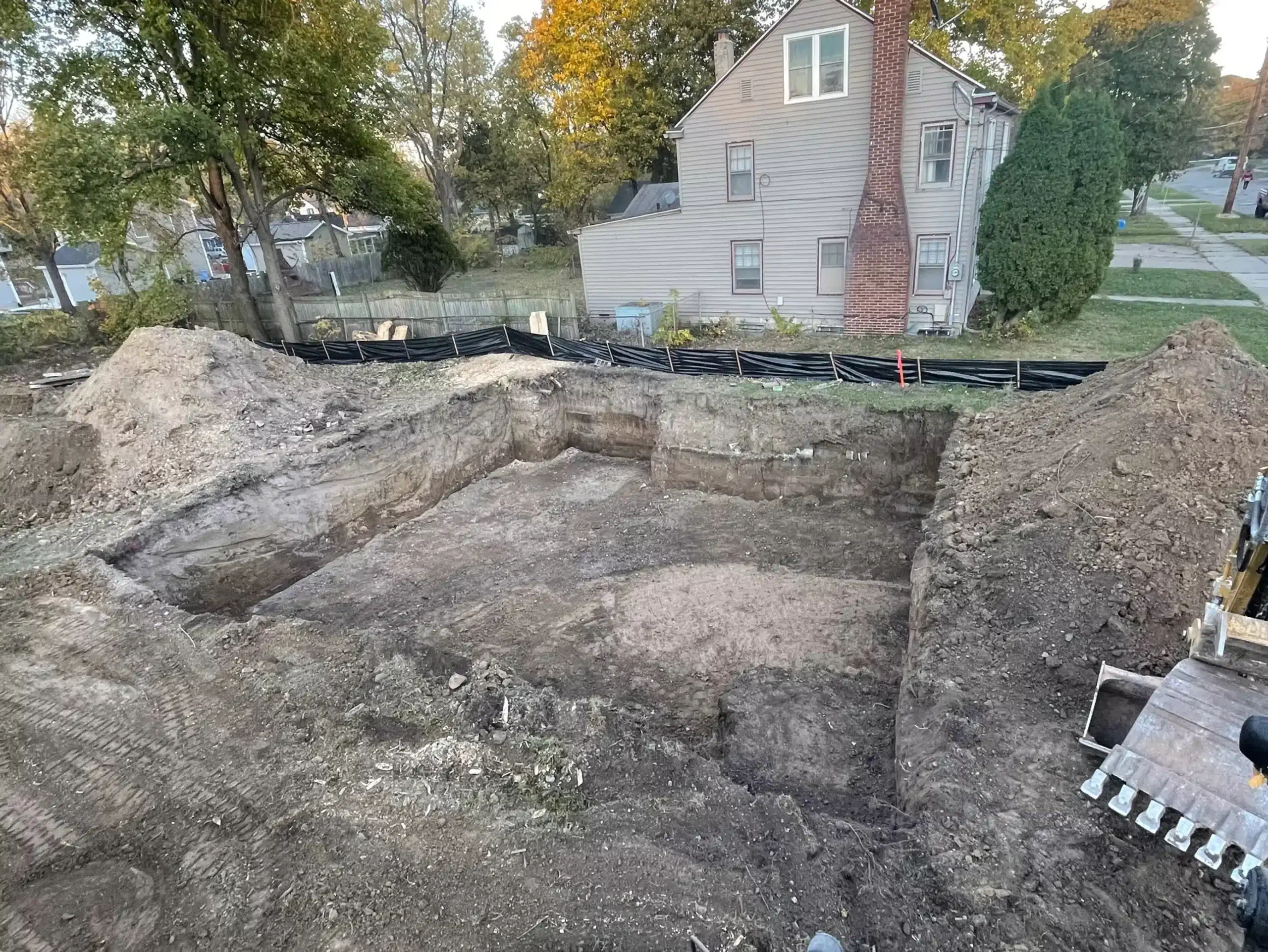 A large rectangular hole has been excavated in the ground at a residential lot by excavation contractors DeWitt MI, with surrounding dirt piles and a house visible in the background. Kenneth Bros