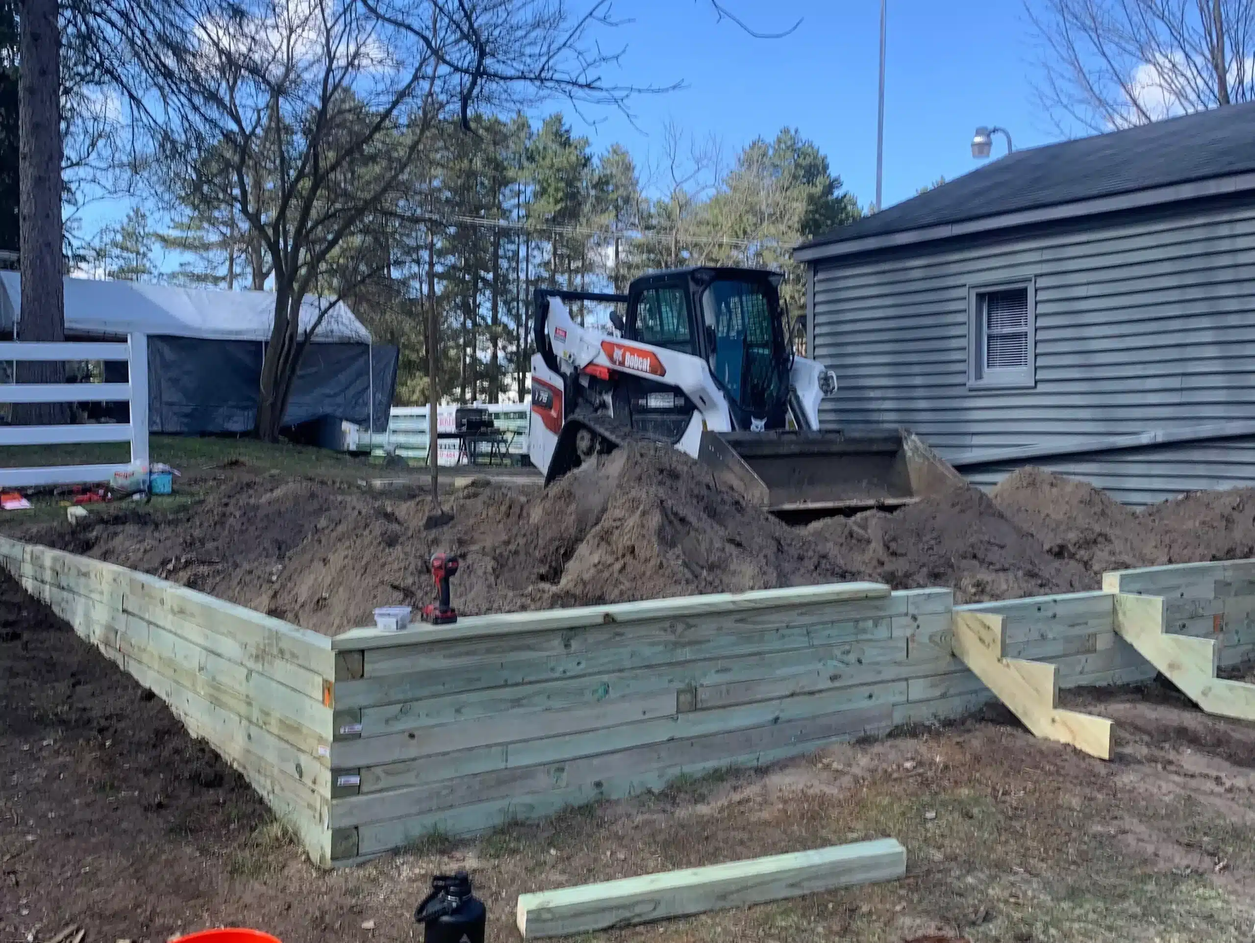 A skid steer loader moves dirt inside a partially built wooden retaining wall frame near a house, as excavation contractors DeWitt MI handle site preparation and grading services amid scattered tools and construction materials. Kenneth Bros