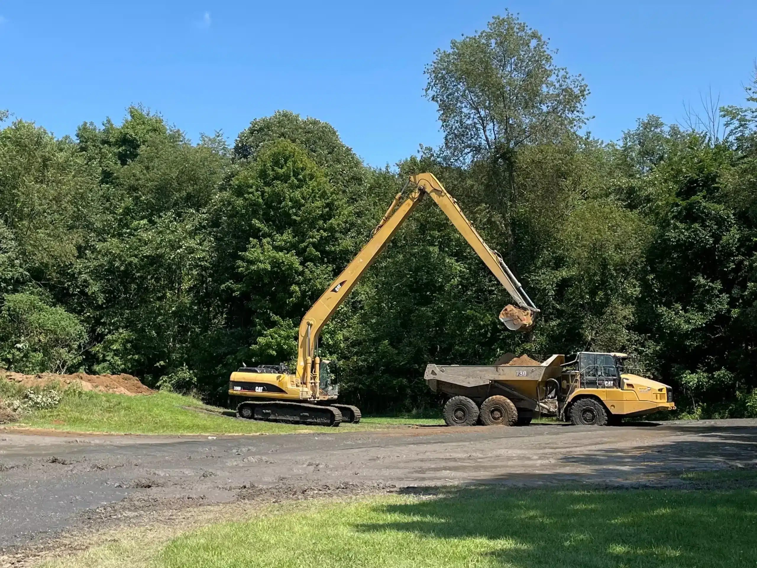 A yellow excavator loads dirt into a dump truck near a grassy field and trees under a blue sky—ideal work for a commercial demolition contractor in mid Michigan or an underground utility installation contractor MI. Kenneth Bros