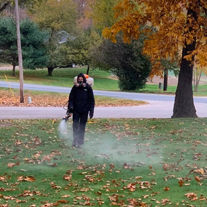 One of our Pest Pros technicians, dressed in protective gear, spraying pest control treatment across a lawn in the fall.