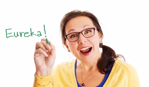 Smiling woman with glasses writes 'Eureka!' in green marker on white background.