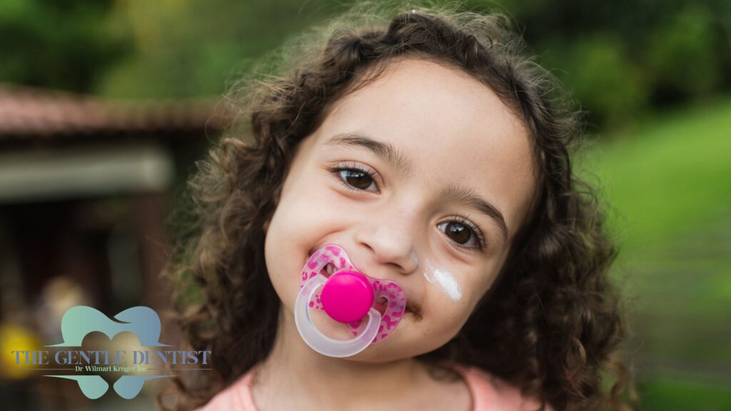 Baby Teeth Questions: Whatโs Normal, Whatโs Not, and What to Do About It 2 Young child with curly hair using a pink dummy, outdoors, showing toddler pacifier and dental habits.