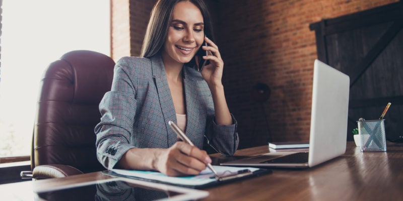 Businesswoman managing prospect lists on phone and laptop in modern office.
