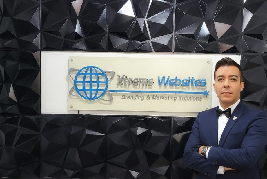 Robert w Xtreme Background Robert Diaz - CEO of Xtreme Websites, stands with arms crossed before a black geometric wall and a sign that reads Xtreme Websites Branding & Marketing Solutions, a leader among top B2B companies, featuring a blue globe logo.