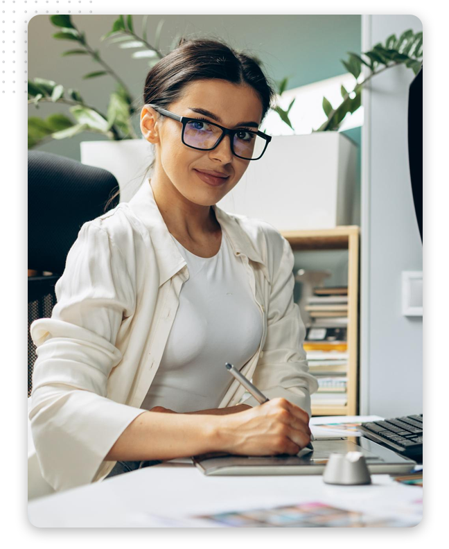graphic-design-process-feat-img A woman wearing glasses and a white shirt sits at a desk, smiling and holding a stylus pen, working on paperwork and a graphic tablet—surrounded by plants and shelves, she appears focused on digital design projects.