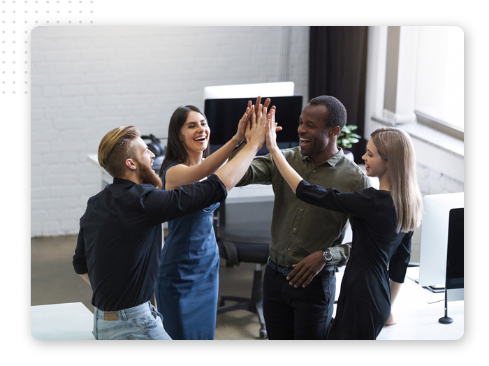 positive-business-meeting-img Four people stand in an office, smiling and giving each other a group high-five, celebrating together. Computers and office furniture are visible in the background.