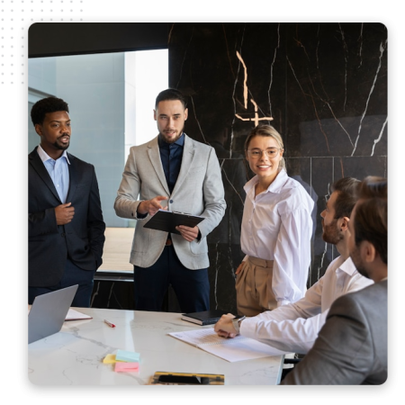 Automator-Enterprise-biz-Img Five people in business attire are having a discussion around a conference table in a modern office, collaborating on strategies for the best CRM software. One stands holding a clipboard, while others listen and smile amid sticky notes and papers.