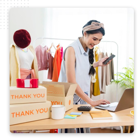 Automator-eCommerce-Img A woman stands at a desk with a laptop, smiling while holding a phone. Around her are thank you boxes, tape, a coffee mug, and clothing on a rack, suggesting she uses the best CRM software to run her online shop from home.