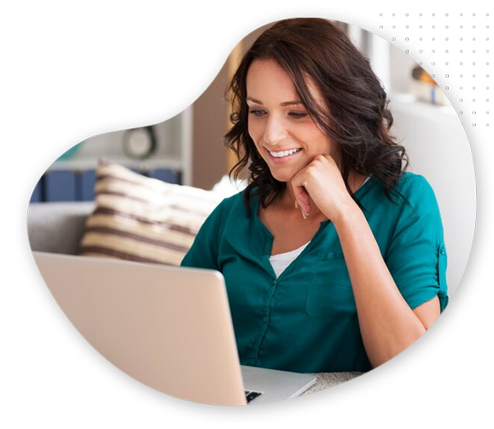 Blog-Home-image A woman with dark hair, wearing a teal blouse, sits at a table and smiles while looking at a laptop screen in a bright, cozy room with striped pillows in the background.